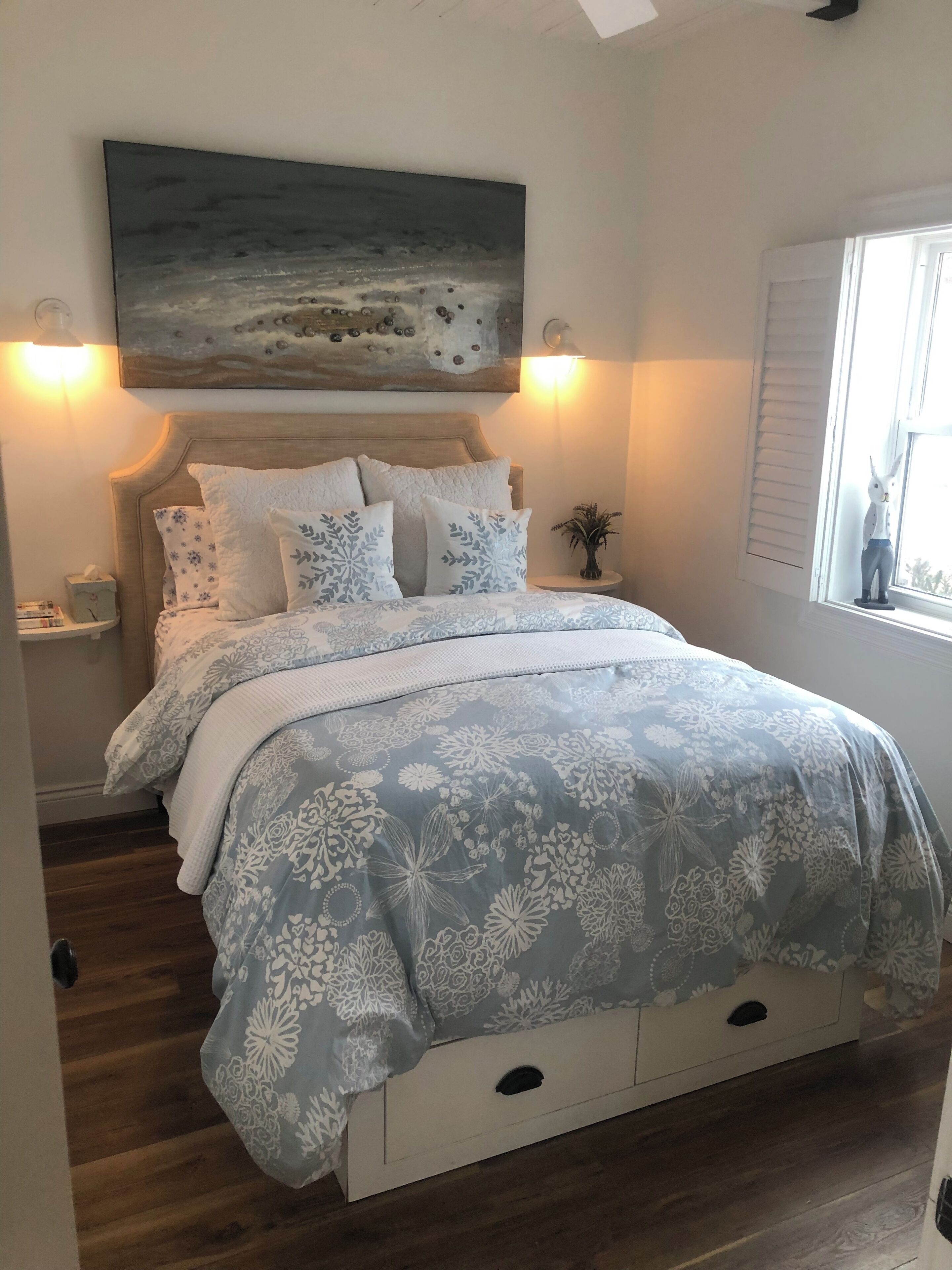 A bedroom with a bed, a nightstand, a lamp, a window, and a painting on the wall.