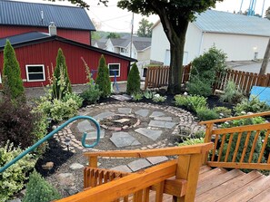 A wooden deck with a stone fire pit surrounded by a garden with various plants and a red building in the background. - The Chestnut Barn built in 1830 is nestled in the quaint Amish town of Winesburg (Winesburg)
