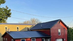 Une grange rouge avec un toit en métal, un bâtiment jaune en arrière-plan et un ciel dégagé.