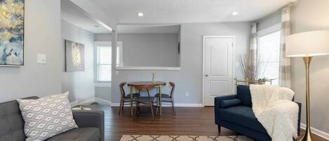 Una sala de estar moderna con un sofá gris, un sillón azul, una mesa de centro redonda y un comedor con mesa y sillas.