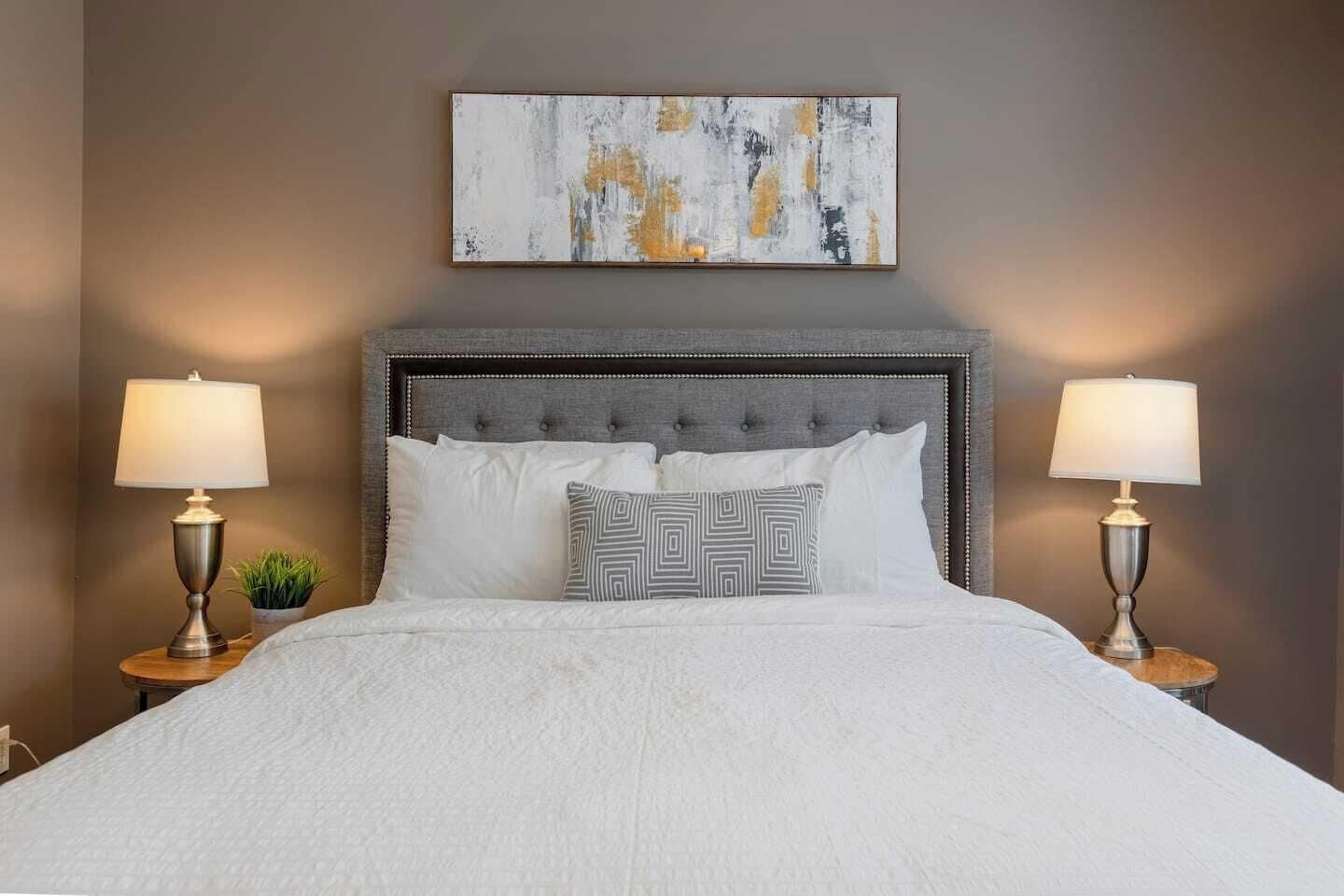 Un letto rifatto con cura, con una testiera grigia rivestita in tessuto, due abat-jour e un cuscino decorativo.
