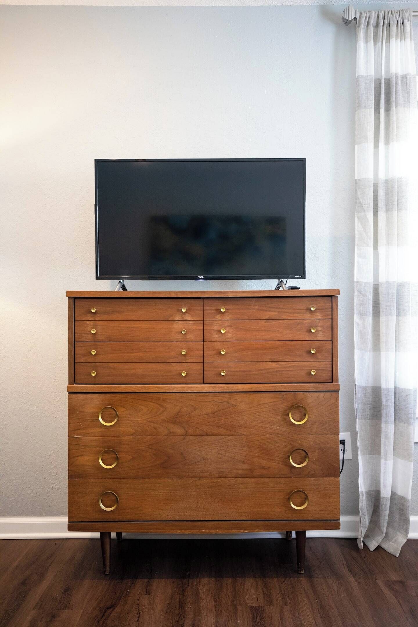 A wooden dresser with a flat-screen TV on top, positioned against a wall with a curtain.