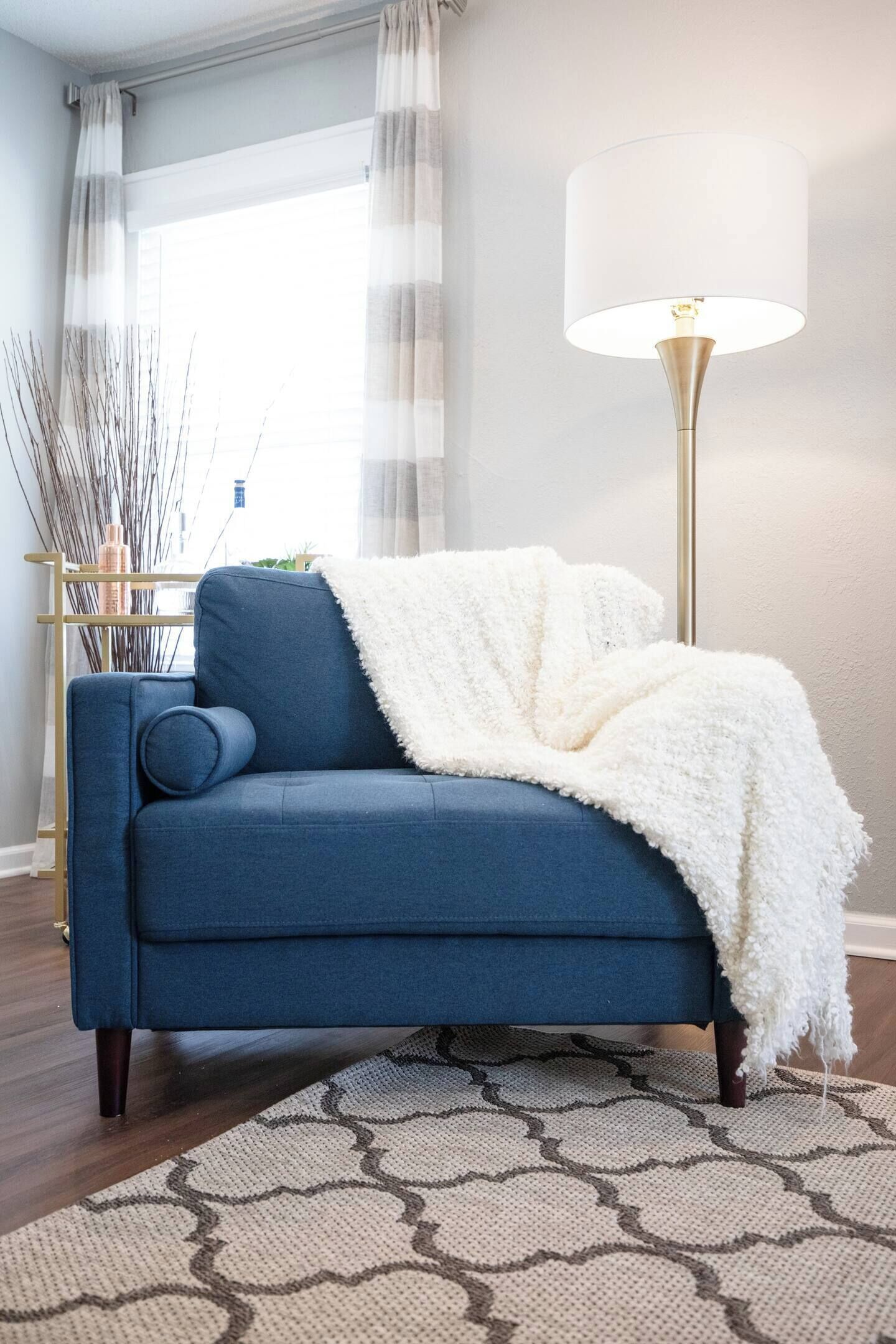 Un canapé bleu avec un plaid blanc, une lampe sur pied et un tapis à motifs dans une pièce au plancher en bois et dotée d’une fenêtre avec des rideaux.