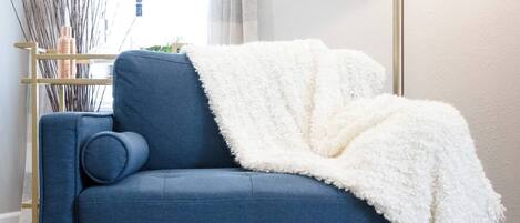 A blue sofa with a white throw blanket, a floor lamp, and a patterned rug in a room with wooden flooring and a window with curtains.