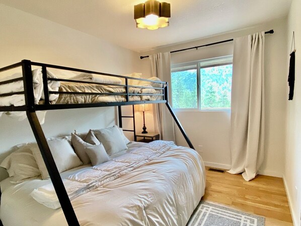 Un lit superposé avec une literie blanche, une table de chevet avec une lampe, une fenêtre avec des rideaux et un plancher en bois.
