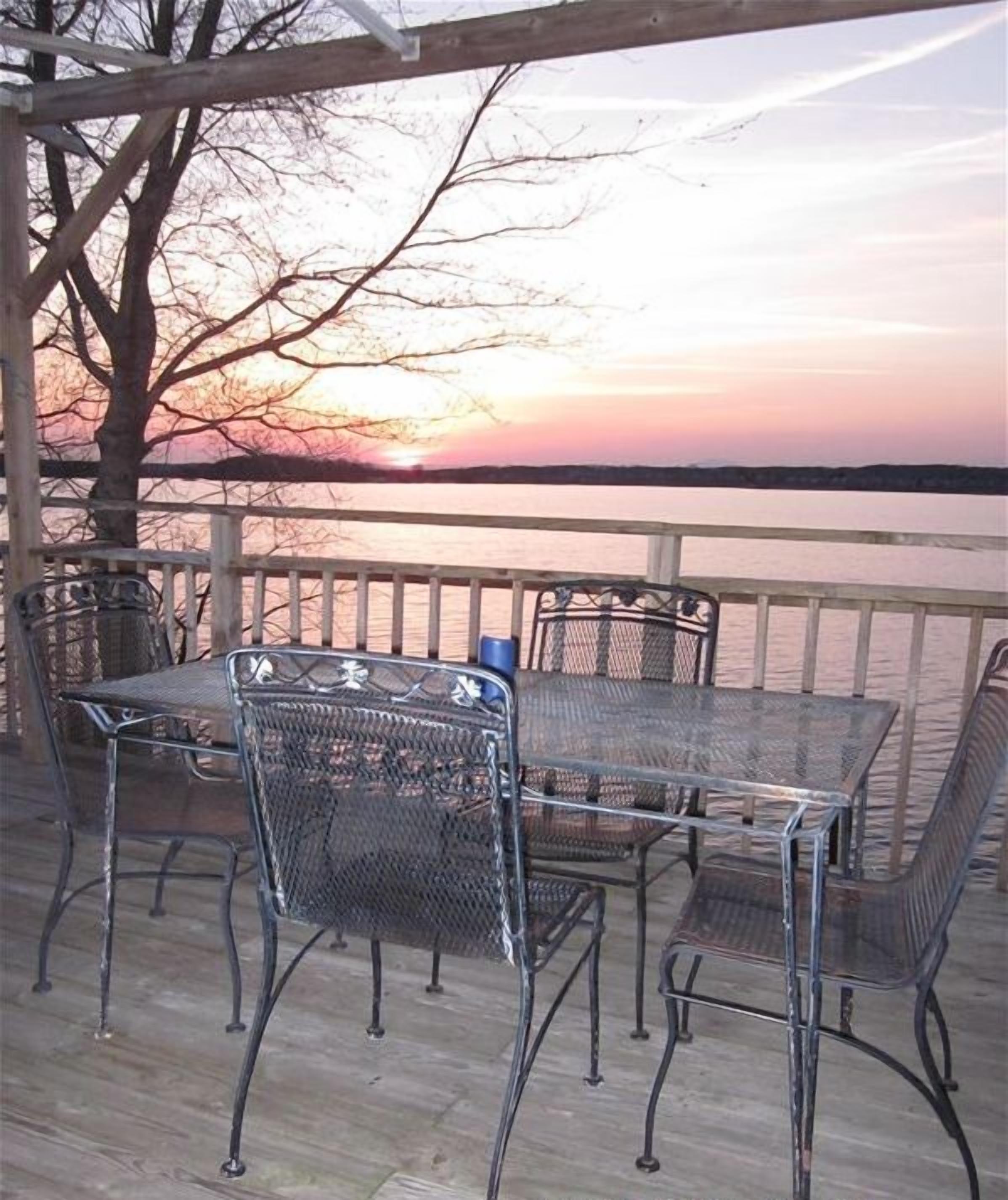 Un espace extérieur aménagé avec des chaises et une table en métal, protégé par un auvent, et offrant une vue sur une étendue d’eau au coucher du soleil.