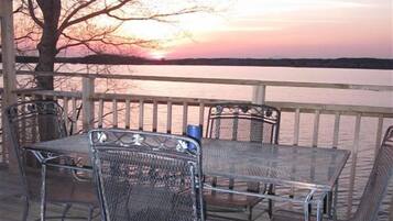 Un espace extérieur aménagé avec des chaises et une table en métal, protégé par un auvent, et offrant une vue sur une étendue d’eau au coucher du soleil.