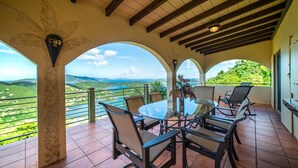Un patio cubierto con una mesa y sillas de vidrio, con vistas a colinas verdes y un cielo despejado.