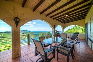 A covered patio with a glass table and chairs, offering a view of green hills and a clear sky.
