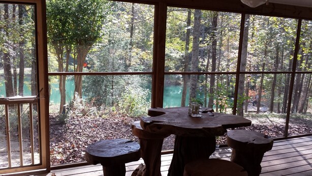 Una mesa de madera rústica con un vaso encima, sobre una terraza con vistas a un bosque y un lago.