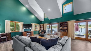Amplia sala de estar con un sofá grande de color gris, un televisor de pantalla plana y una estantería. Amplias ventanas ofrecen vistas al exterior.