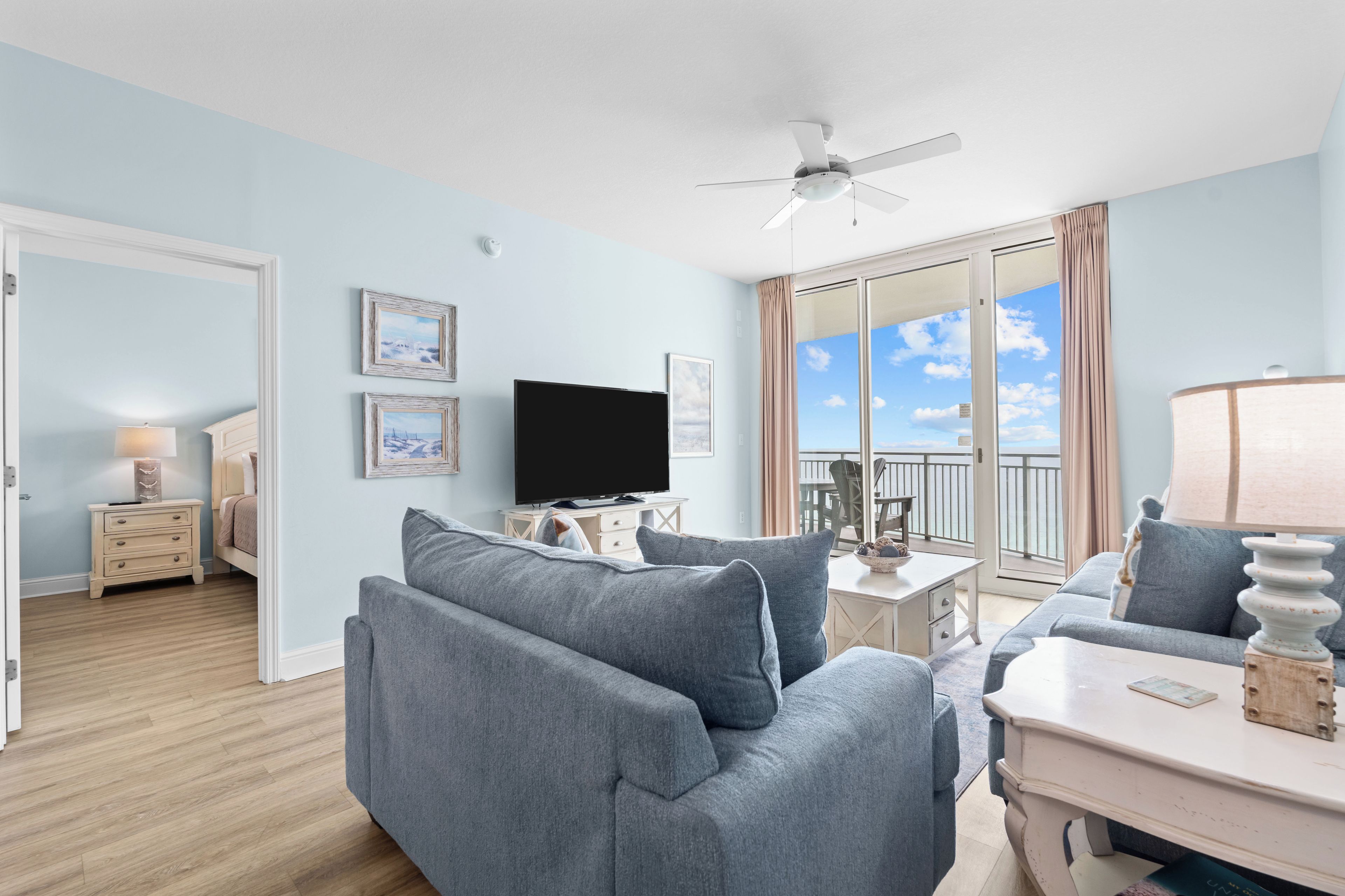 A modern living room with a flat-screen TV, grey sofas, a ceiling fan, and a balcony with a view.