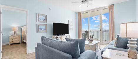 A modern living room with a flat-screen TV, grey sofas, a ceiling fan, and a balcony with a view.