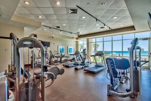 Une salle de sport équipée de divers appareils d’exercice, dotée de grandes fenêtres et offrant une vue sur la plage.