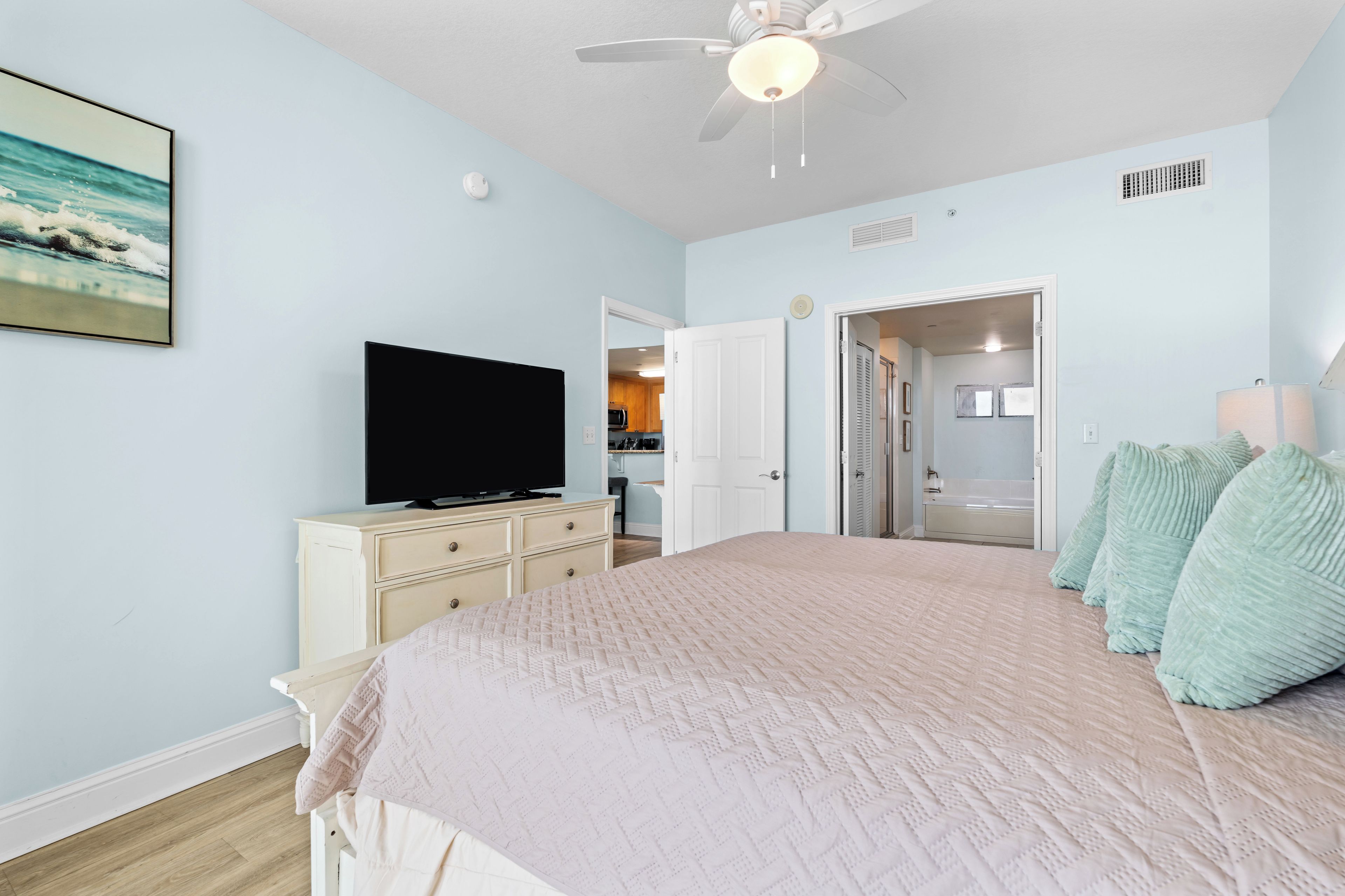 Un dormitorio con cama, televisión, ventilador de techo y puerta que da a un baño.