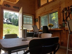 Interior de uma cabana de madeira com uma mesa de jantar, duas cadeiras, uma janela com vista para uma casa e vegetação, e uma parede decorada com um peixe.