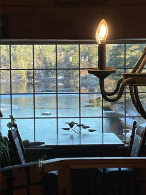 Un comedor con una mesa y sillas, una vela sobre un atril, y vistas a un cuerpo de agua y árboles a través de amplios ventanales.