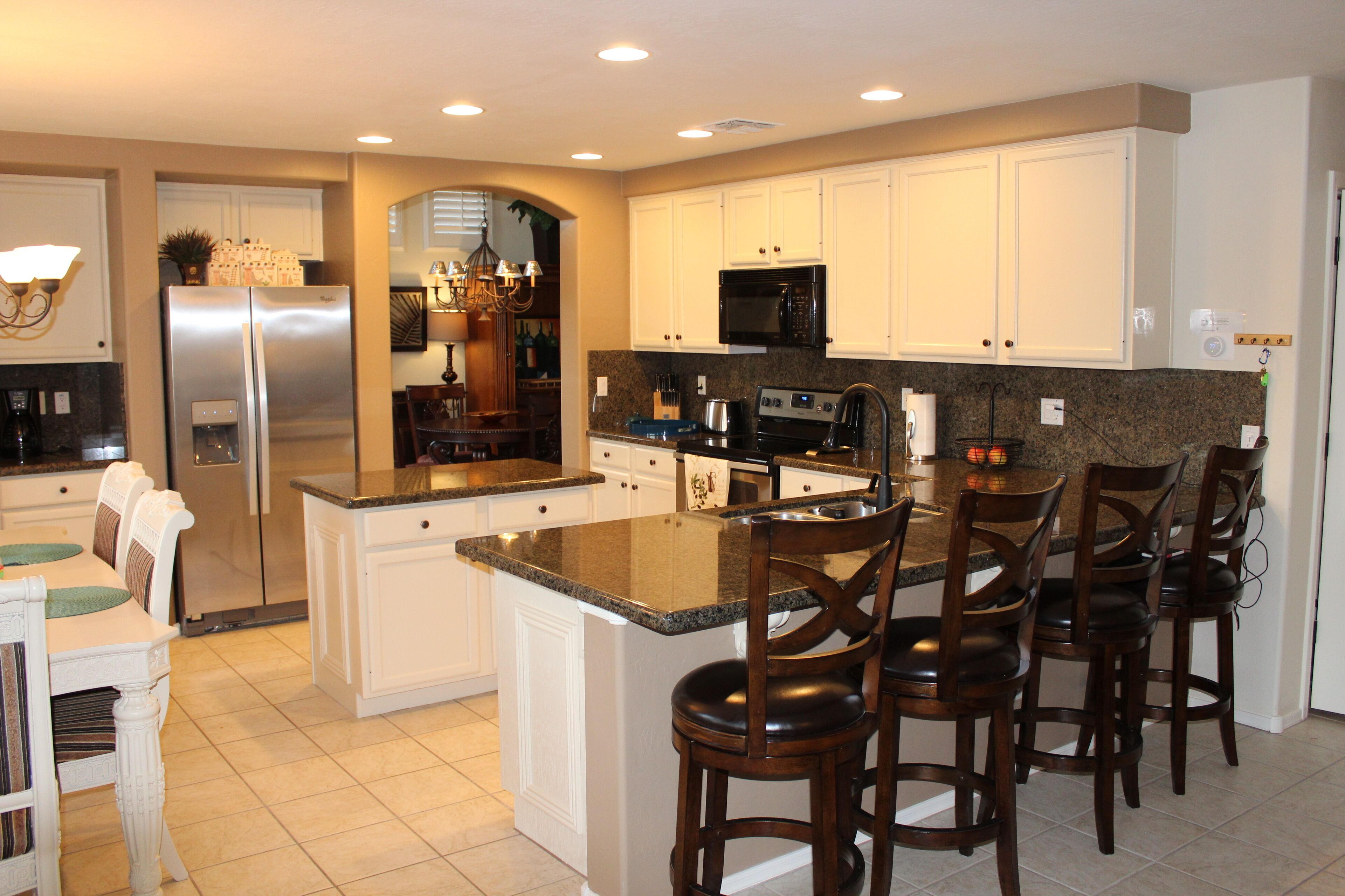 A spacious kitchen with white cabinets, a stainless steel refrigerator, a granite countertop, and a bar area with four stools.