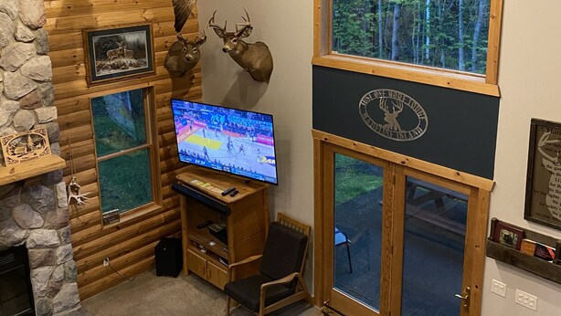 Interior acogedor de una cabaña con chimenea de piedra, venados montados como adorno, un televisor y un ventanal con vistas a un bosque.