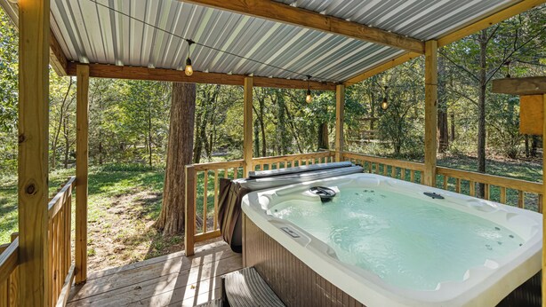 A hot tub under a wooden deck with a metal roof, surrounded by trees. - The Magnolia — A River-View Bungalow with Hot Tub (Pencil Bluff)