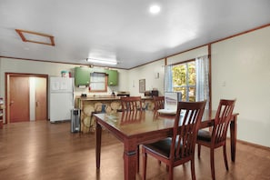 A dining area with a wooden table and chairs, a kitchen with green cabinets, and a window with curtains. - The Magnolia — A River-View Bungalow with Hot Tub (Pencil Bluff)