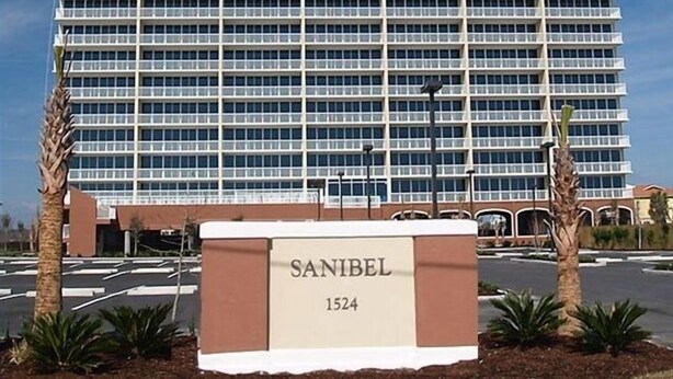 Un immeuble de grande hauteur avec une façade constituée d’une grille de fenêtres, une enseigne indiquant « SANIBEL 1524 » et un parking bordé de palmiers.
