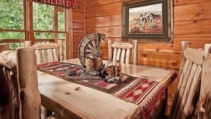 Interior de una cabaña de madera con una mesa de comedor, sillas, una rueda decorativa y un cuadro enmarcado con caballos.