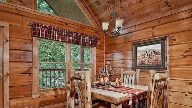 Interior de una cabaña de madera con una mesa de comedor, sillas, una araña y una ventana adornada con un toldo escocés.