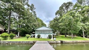 A wooden dock leading to a house with a glass facade, surrounded by lush greenery and a calm water body.