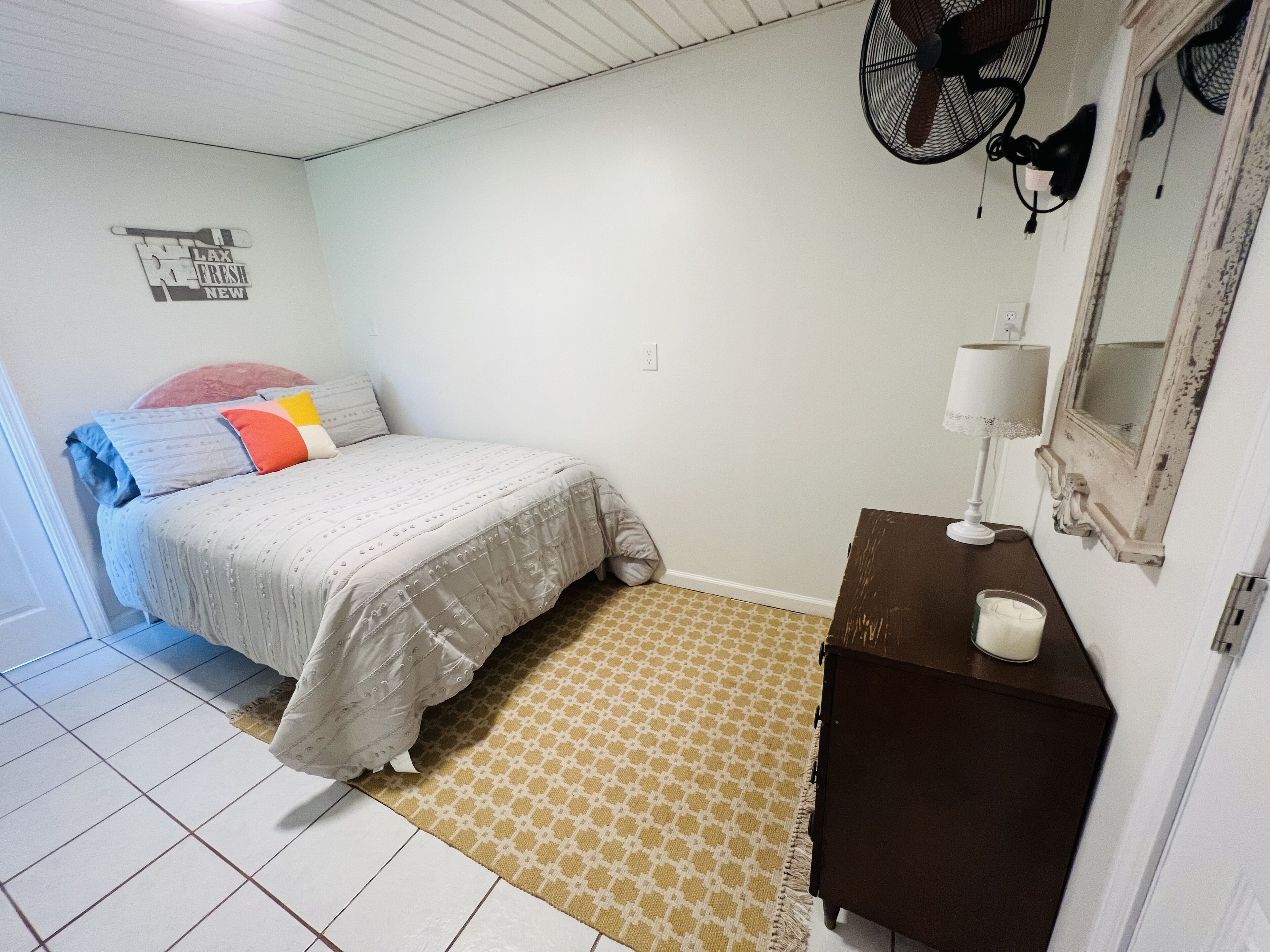 Een slaapkamer met een bed, nachtkastje met lamp, spiegel, wandventilator en een kamerbreed tapijt met patroon.