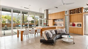 A modern kitchen with a central island, a dining area with a white table and chairs, and a living area with a black leather sofa and a coffee table.
