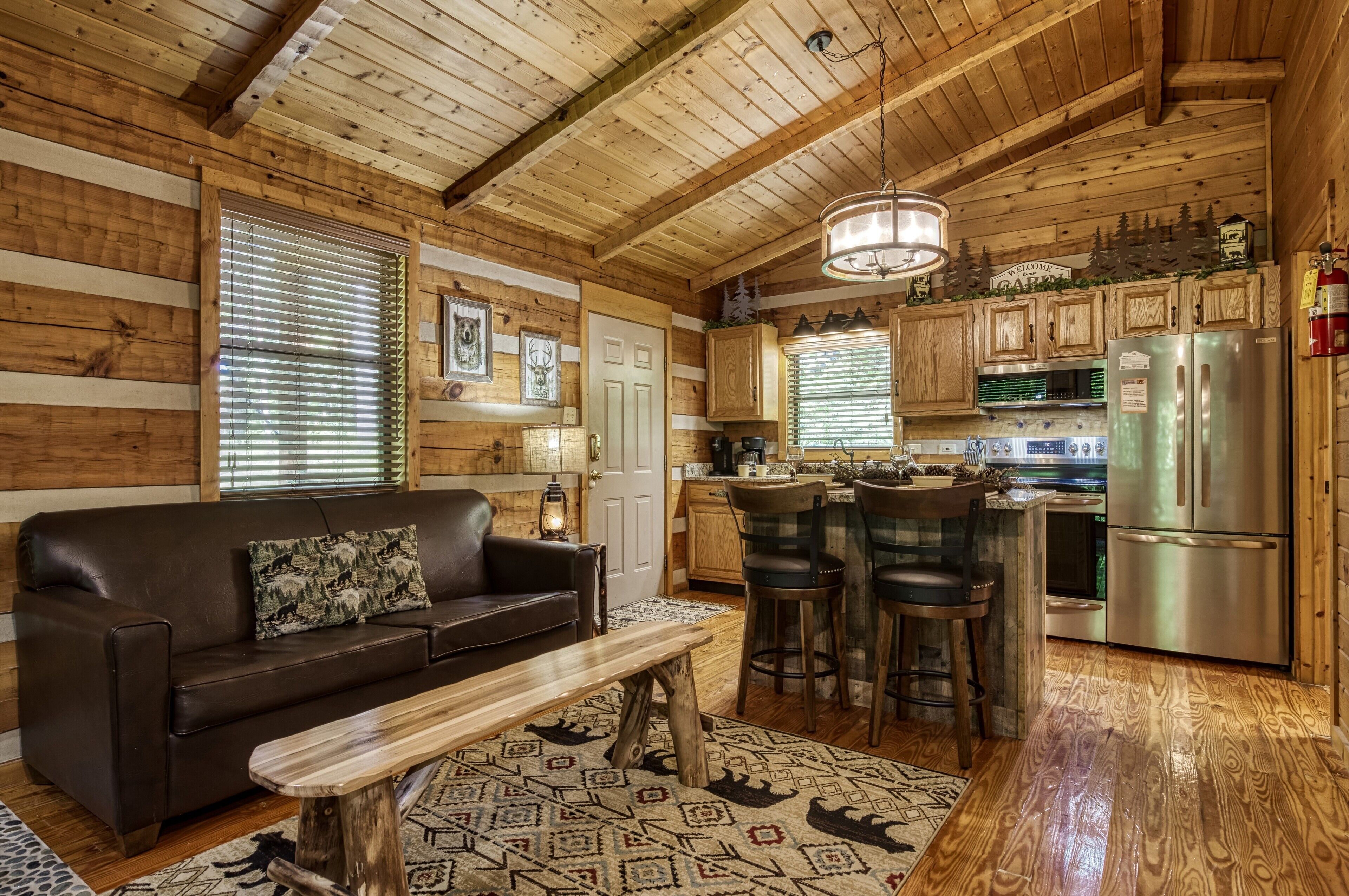 Acogedor interior de cabaña con detalles de madera, un sofá de cuero, zona de comedor y cocina con refrigerador de acero inoxidable.