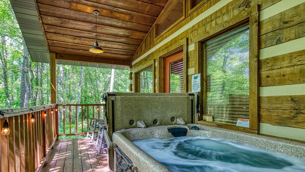 A wooden cabin with a hot tub, wooden deck, and large windows overlooking a forest. - Secluded Studio Cabin Rental-Honeymoon/Anniversary Getaway-dog Friendly (Pigeon Forge)