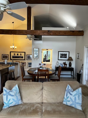 Uma sala de estar ampla, com um sofá bege, mesa de jantar de madeira e cadeiras. Uma cozinha visível ao fundo, com uma janela e vista para o exterior.