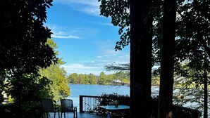 Eine Holzterrasse mit Stühlen und einem Tisch, mit Blick auf einen See, Bäume und ein Haus in der Ferne.
