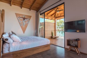 Ein modernes Schlafzimmer mit einem großen Bett, einer Holzbalkendecke, einer Schiebeglasdoor zum Balkon und einem an der Wand montierten Fernseher.