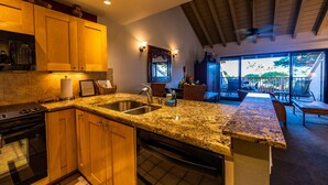 A kitchen with granite countertops, wooden cabinets, a stainless steel sink, and a dishwasher. There is a dining area with a table and chairs visible through a glass door. - Beautiful 2 Bedroom Oceanview Condo at Maui Kamaole The Perfect Maui Escape! (Kihei)