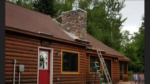 Hirsimökki, jossa on kivinen savupiippu, punainen ovi ja taloa vasten nojaava tikapuu.