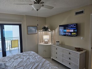 Een hotelkamer met een bed, een dressoir, een televisie waarop de OXC TV-zender te zien is, een plafondventilator en uitzicht op de oceaan door een deur.