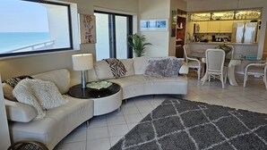 Ein Wohnzimmer mit einer gebogenen Couch, einem kleinen Tisch mit einer Lampe, einem gemusterten Teppich und Blick auf den Ozean durch große Fenster.