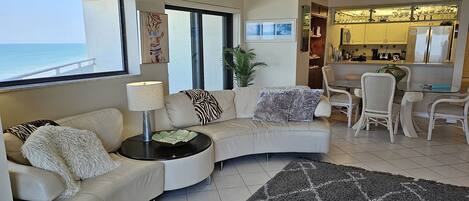 A living room with a curved sofa, a small table with a lamp, a patterned rug, and a view of the ocean through large windows.