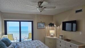 Ein Schlafzimmer mit einem Bett, einem Deckenventilator, einem Fernseher, einem Schminktisch und Blick auf den Ozean durch eine Balkontür.