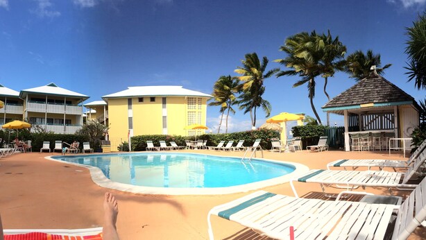 A pool area with lounge chairs and umbrellas, surrounded by palm trees and a building with a yellow facade. - Beachfront Caribbean Paradise at Colony Cove (Christiansted)