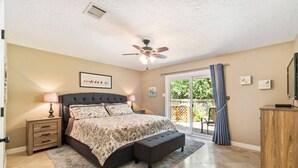 Une chambre à coucher comprenant un lit, des tables de chevet, un ventilateur de plafond, une porte-fenêtre coulissante donnant sur un balcon et une armoire en bois.