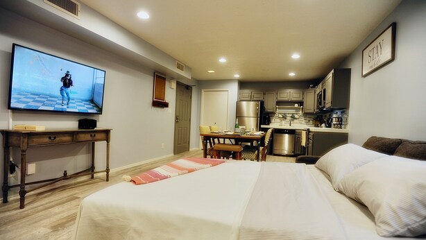A bedroom with a large bed, a desk with a TV, a kitchenette, and a dining area.