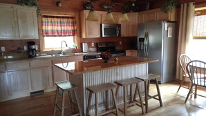 A kitchen with wooden cabinets, a stainless steel refrigerator, a microwave, and a sink. There are two stools and a window with curtains. -  Secluded Mountainside Cabin, Close to Town, No 4 wheel drive needed!  Hot Tub! (Bryson City)