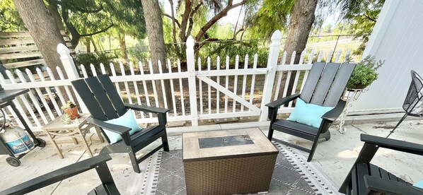An outdoor patio with two black chairs and a small table, surrounded by a white picket fence and trees.
