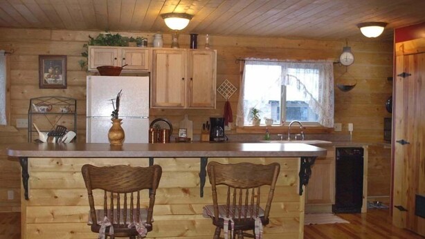 Een keuken in chaletstijl met houten wanden, een houten bar, twee houten barkrukken, een koelkast, een spoelbak en een raam met gordijnen.