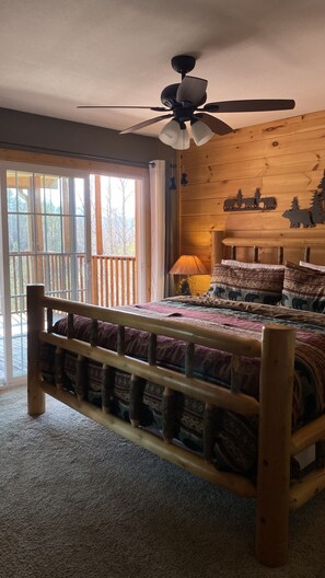 A wooden-framed bed with a patterned bedspread, a ceiling fan, and a window with a view of trees.
