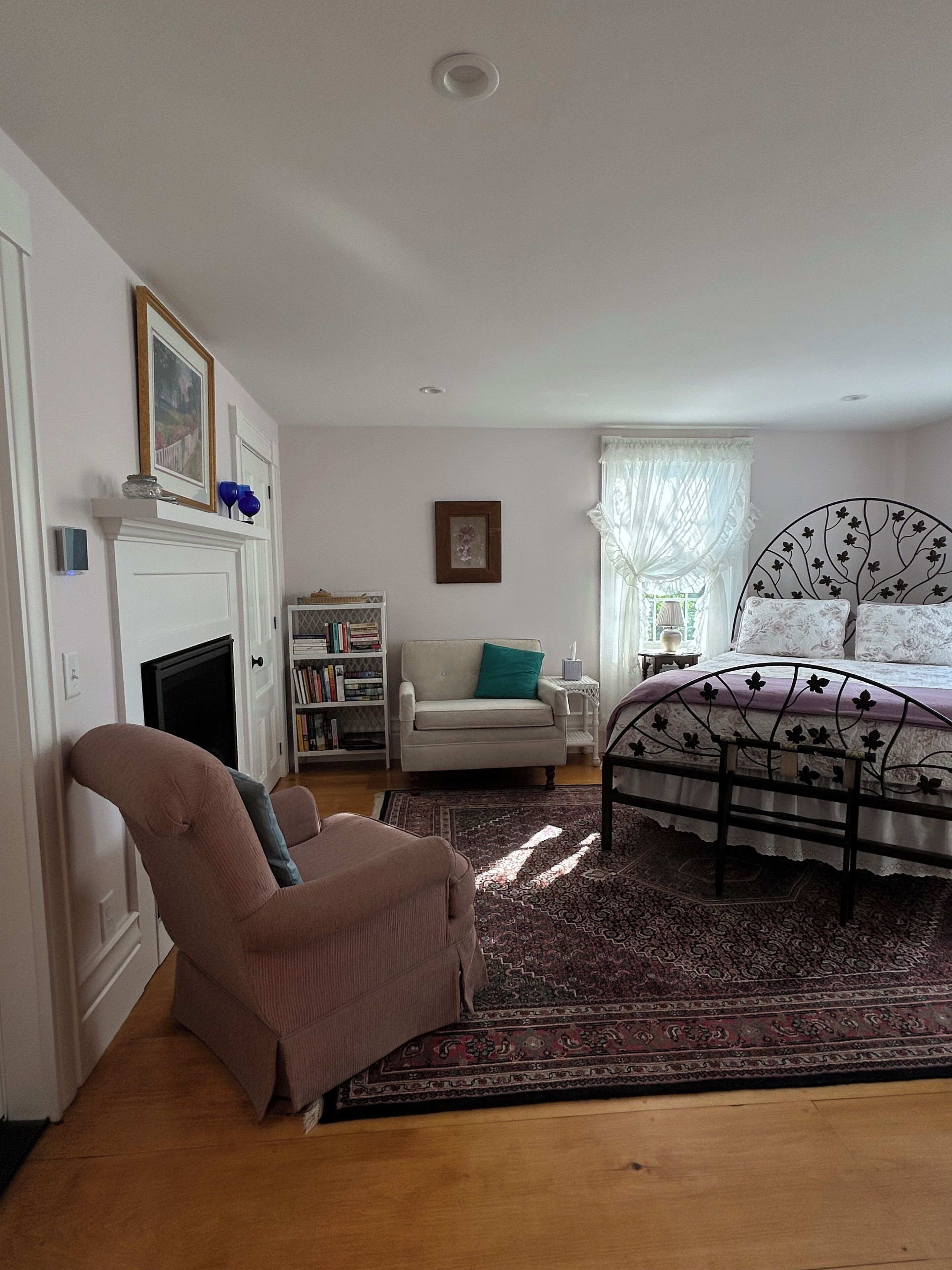 Quarto com cama, cadeira, lareira, estante de livros e janela com cortinas.